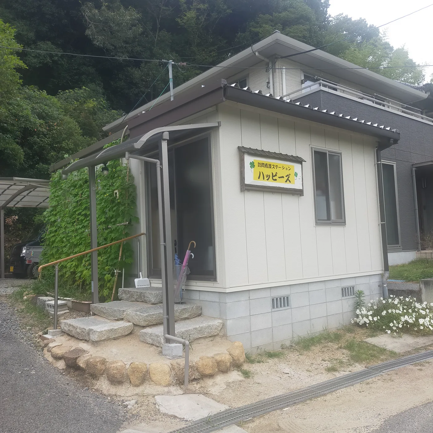 訪問看護ステーションハッピーズの事業所の外観。白い壁の平屋に木枠の看板がかかり、入口の階段に手すりがある