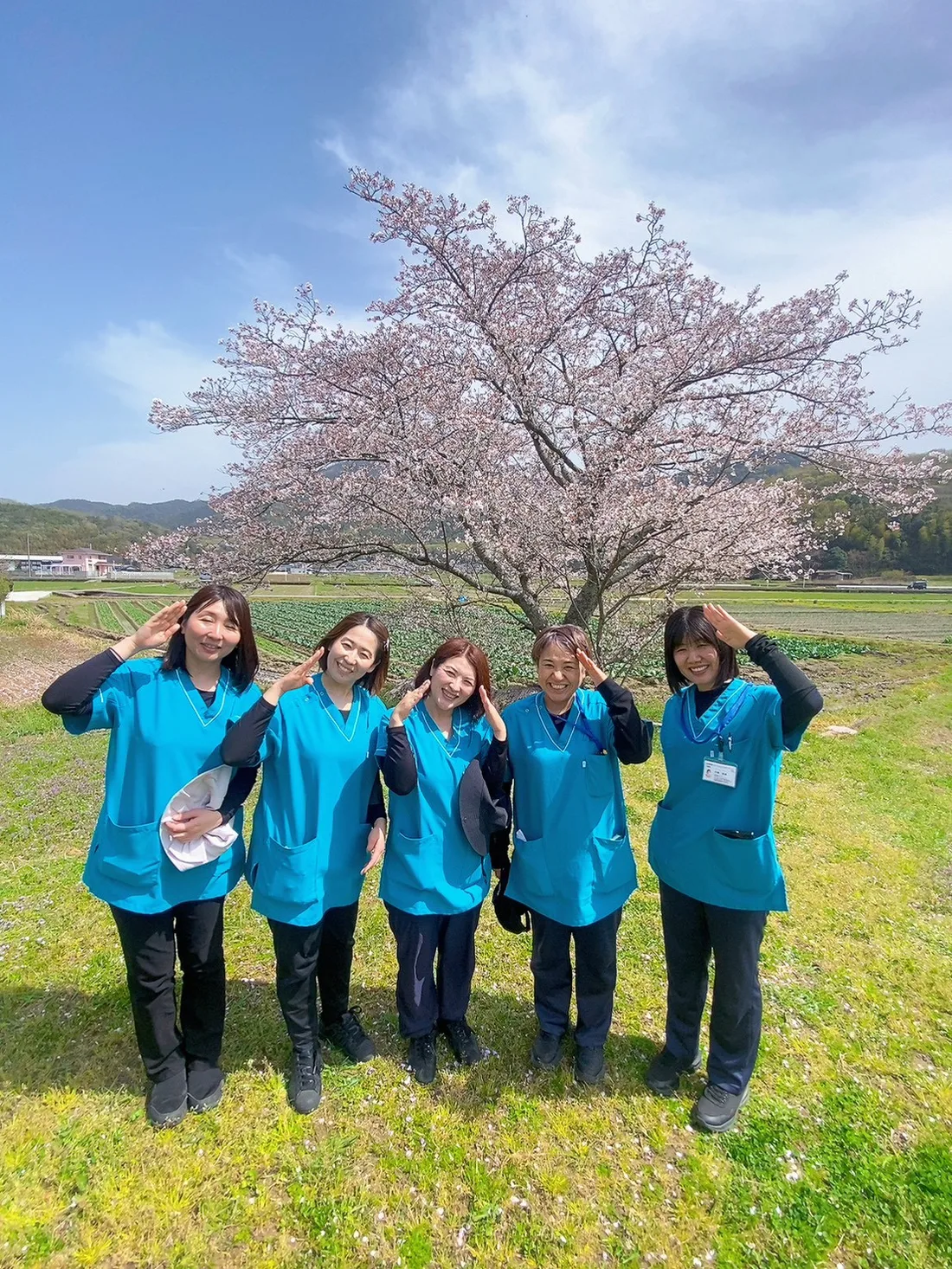 満開の桜の前で敬礼ポーズをとるハッピーズのスタッフ5人